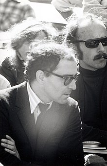 Photo noir et blanc d'un homme de 38 ans, costume noir, cheveux bouclés, lunettes noires, de profil dans une foule