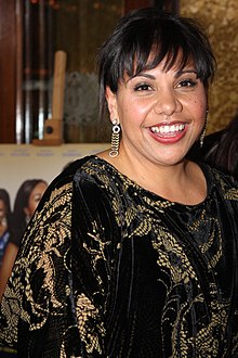 A dark-haired, smiling woman with long, dangling earrings looks right toward the camera