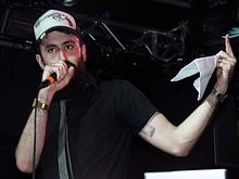 Image of the white, bearded rapper performing on stage, holding a microphone. He is wearing a black t-shirt, a grey tie and a "trucker cap". The cap has a pistol morphed into a microphone.