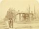 Sepia-toned 1890s photograph showing two men in late 19th-century clothing standing on the street corner in front of a brick house