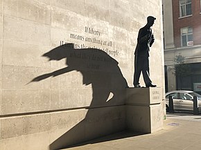 statue en pied d'un personnage vêtu d'un costume, sur un socle contre un mur