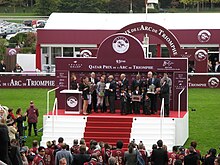 Distant photo of a podium with several people. In front of it are photographers.