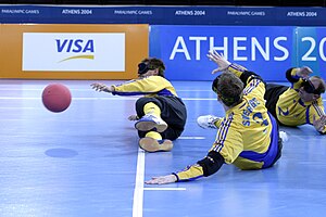 Três atletas, usando vendas, estão deitados na quadra, durante uma partida de goalball.