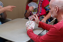 An einem kleinen grauen Tisch sitzen drei Personen, die man jeweils nur teilweise sieht. Die Person im Vordergrund hat zehn Spielkarten in der linken Hand und fügt mit der rechten zwei weitere hinzu. Eine Person im Hintergrund hält zehn Spielkarten verdeckt in der rechten Hand. Von der dritten Person am rechten Bildrand sieht man nur eine Hand, die in die Bildmitte zeigt.