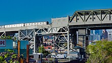 Grauer Stahlviadukt, graue hohe Fachwerkbrücke, darauf silbriger U-Bahn-Zug. Im Hintergrund bunter eine Stadtlandschaft mit verschieden hohen Gebäuden, in verschiedenen Farbtönen und jeweils nur ausschnittweise zu sehen. Vorne Vegetation und ebenfalls leicht farbige Industriebauten.