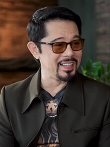 Christopher de Leon wearing a black suit jacket and a print shirt, with tinted glasses, grinning and looking left of the camera