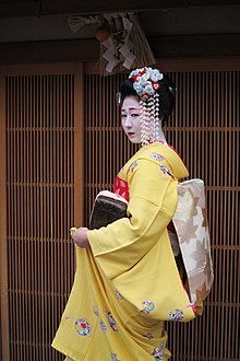 A young apprentice geisha stood outside a traditional Japanese building. Her yellow kimono's sleeves are shorter than a maiko's, and she wears more hair accessories than a maiko would.