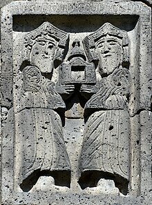 Two bearded figures in stone relief, holding an object that looks like an open door with a bell on top.