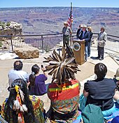 American flag and group of four or five speakers at a podium in front of audience including a Native American in headdress