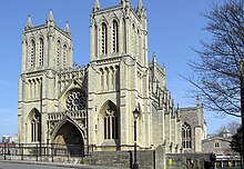 A stone built Victorian Gothic building with two square towers and a central arched entrance underneath a circular ornate window. A Victorian street lamp stands in front of the building and on the right part of a leafless tree, with blue skies behind.