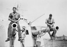 A twin barrelled warship turret. Four young sailors are working on the barrels: two are sitting astride the barrels, while the other two work from the deck.