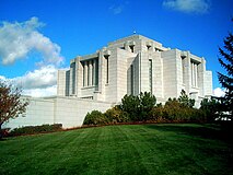 The Cardston Alberta temple sits in the middle of the photo, in the style of a Mayan/Aztec design meshed with the Prairie School architectural styling. A large grassy lawn can be seen in the foreground, along with a blue sky.
