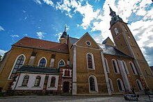 Barevná fotografie s pohledem na hlavní loď kostela, hranolovitou věž a přístavby v podobě kaplí