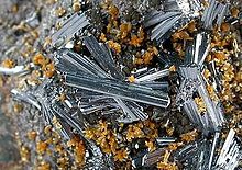 A close view of a rock crusted with groups of glassy, lustrous, silvery-blue hutchinsonite, in tight clusters of loosely aligned needle-like crystals, among smaller clusters of tiny orange-brown crystals