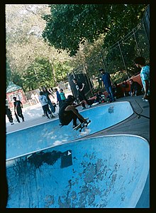 A person does an airborne trick between two light blue bowls in the skate park in Owl's Head. A few people watch from above the bowl.