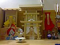 A small pinewood shrine, featuring a plain torii gate decorated with streamers, a small mirror in front of a cup, and a blue daruma doll.