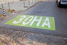 Grün markiertes Rechteck auf einer Straße, darin die mit weißer Farbe die kyrillische Schrift ЗОНА. Vorne erkennt man Kopfsteinpflaster, am Rande der Straße Poller und parkende Autos.