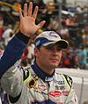 A man in his early thirties waving to the crowd with his right hand and is wearing white and blue racing overalls along with a similarly colored baseball cap