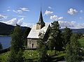 Mala kamnita cerkev z majhnim zvonikom na lesenem stolpu stoji na pokopališču s pogledom na jezero in gore.