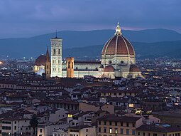 The Florence Cathedral, illuminated at night, showing the large red brick dome, a decorated white marble nave, and a vertical, white marble bell tower to the left. Mountains are visible in the background and a dark, low-lying city in the foreground.