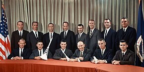 Photographie couleur des aspirants astronautes posant en costume près d'un drapeau des États-Unis. Certains tiennent en main des maquettes de vaisseaux spatiaux.