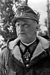 A black-and-white photograph of a man with glasses wearing a military uniform underneath a fur collared coat, field cap and neck order in shape of an Iron Cross.