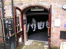 Arched doorway through a weathered red brick wall with sign "Magical Experience" above. On the wall inside the doorway is stylised silhouette of the four Beatles members