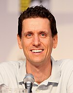 A man with brown hair who is slightly crouched forward and speaking into a microphone.