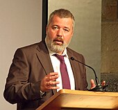 A man with a full beard, Dmitry Muratov, speaking at a podium