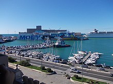 Vue d'un port avec au premier plan une voie de circulation de voitures, au second plan des bateaux de plaisance, et au fond la mer et un volumineux ferry