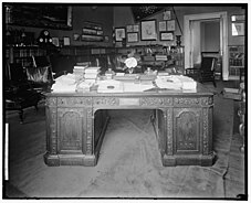 A black and white image of the Resolute desk in a room with dark walls, low bookcases, and memorabilia displayed on the walls. The desk is cluttered with stacks of paper, objects, and a large flower.