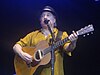 A man behind a microphone holding an acoustic guitar and wearing a yellow shirt and black hat.