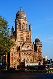 A brown building with three domes and a triangular facade. A tree and a bus on either side of it