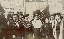 A group of suffragist woman standing together and talking, with banners in the background