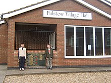 Frontage of a brick-built hall with two young Fulstowians standing by the entrance and the village's war memorial.