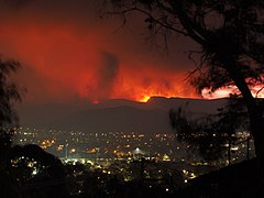 Photograph of evening in a valley settlement. The skyline in the hills beyond is lit up red from the fires.