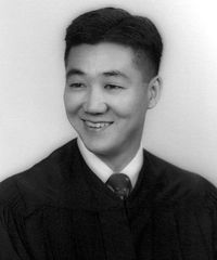 Head and shoulders of a young man with a bright smile and neatly combed hair wearing what appears to be a graduation gown over a shirt and tie.