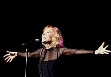 A smiling woman with short blond hair faces a microphone. Her arms are flung wide open to either side.