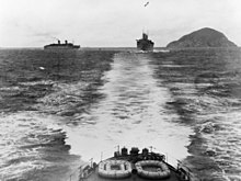 Photograph of the stern and wake of a ship. Two more ships are following in the distance. There is a rocky island to the right.