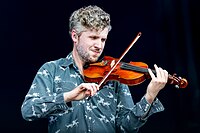 Upper body shot of a man playing a violin nestled under the left side of his chin. He wears a blue shirt with small, white cowboys riding horses as they rope long-horned cattle. The man has fair to brown hair and beard.