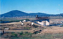 Kings Avenue Bridge under construction. The bridge is incomplete and only the pylons and platform on the southern side are in place. The site is surrounded by dust and the lakebed is dried out.