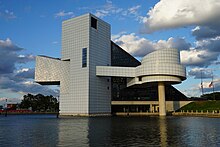 A gray tiled building rises over a lake, with a cylinder set on a narrow pole, and a sloping glass wall on one end.