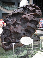 Sebuah batu besar berbentuk telur berwarna hitam dengan struktur berpori berdiri di atasnya, miring