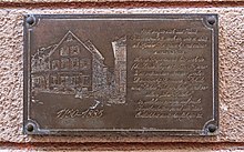 Sur la plaque figure une gravure de maison, à droite un texte descriptif.