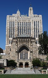 An imposing multi-story Gothic Revival building rises above a green lawn toward a deep blue sky. The building is highly decorated with stonework, heavy wooden entrance doors, and vertical columns of stained-glass windows.