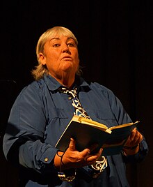 Cathie Dunsford ist im dämmrigen Licht ein Buch haltend zu sehen. Sie hat halblanges helles Haar und trägt ein dunkelblaues Oberteil.