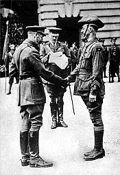 Two men in military uniform smiling and shaking hands. The man on the left is older and slightly stooped. People are gathered in the background.