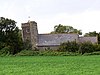 Low building with tiled roof and non-vertical square tower, surrounded by trees and with grass in the foreground.