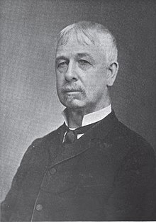 A white-haired man wearing a high-collared white shirt, black tie, and black jacket