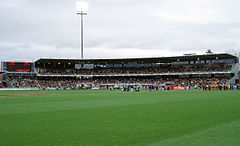 A two tier stand and scoreboard filled with people in the backdrop of an oval grass playing surface.